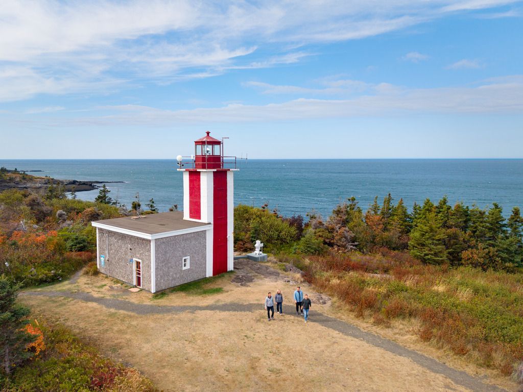 De rood-witte vuurtoren Point Prim Lighthouse nabij Digby, Nova Scotia, aan zee, met een paar mensen op een zandpad onder een blauwe hemel.