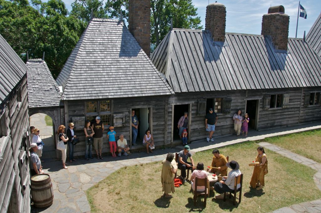 Historisch dorpsgezicht in het Port-Royal National Historic Site te Nova Scotia met rietgedekte huisjes en mensen die buiten rond een tafel zitten.