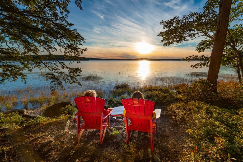 Twee rode stoelen aan een meer in het Kejimkujik National Park op Nova Scotia bij zonsondergang, met bomen langs de oever en gouden licht op het water.