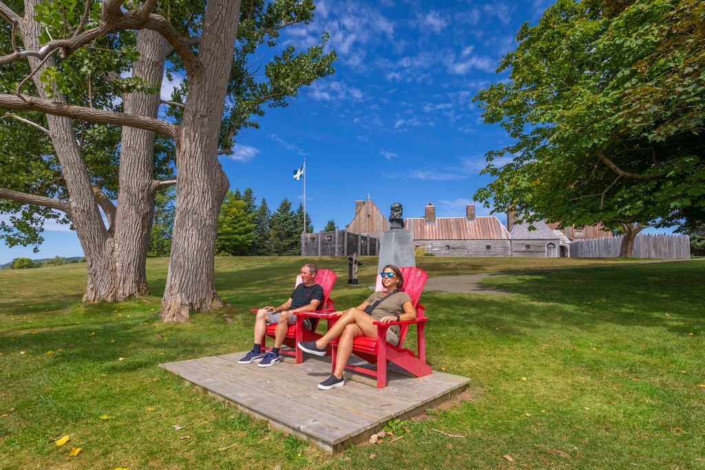 Twee mensen zitten voor het Port-Royal National Historic Site, Nova Scotia, op rode Adirondack-stoelen op een grasveld naast bomen en een stenen muur onder een blauwe hemel.