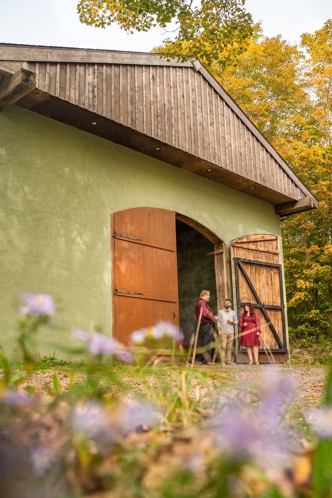 Kind bij een schuurdeur van Casa Nova Fine Beverages in Bear River East, Nova Scotia, naast een houten kar, met bloemen op de voorgrond en herfstbomen op de achtergrond.