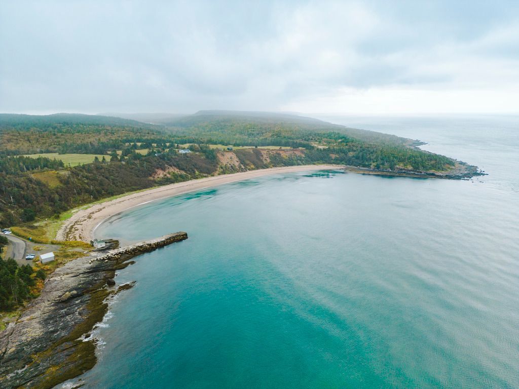 De turquoise baai van Sandy Cove Beach op Nova Scotia met een gebogen zandstrand, rotsachtige kust en groene heuvels onder een bewolkte hemel.