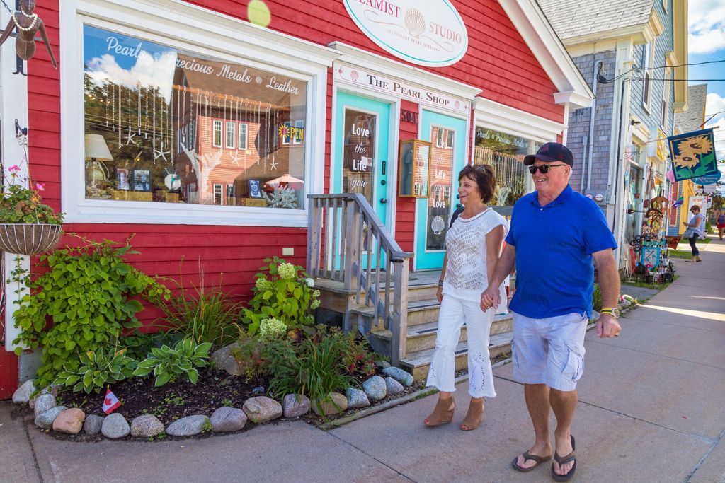 Twee mensen lopen langs een rode winkelpui in een zonnige straat in Mahone Bay, Nova Scotia, met bloembakken en een buitentrap.