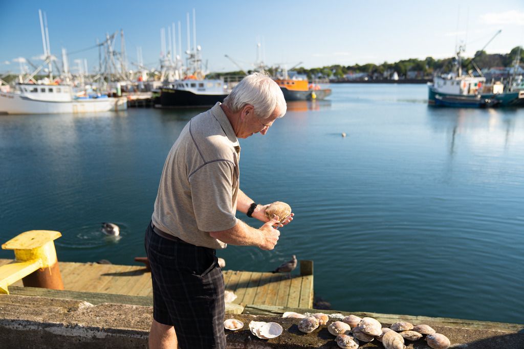 Een visser maakt Sint Jacobsschelpen schoon op de steiger in de haven van Digby op Nova Scotia met boten op de achtergrond.