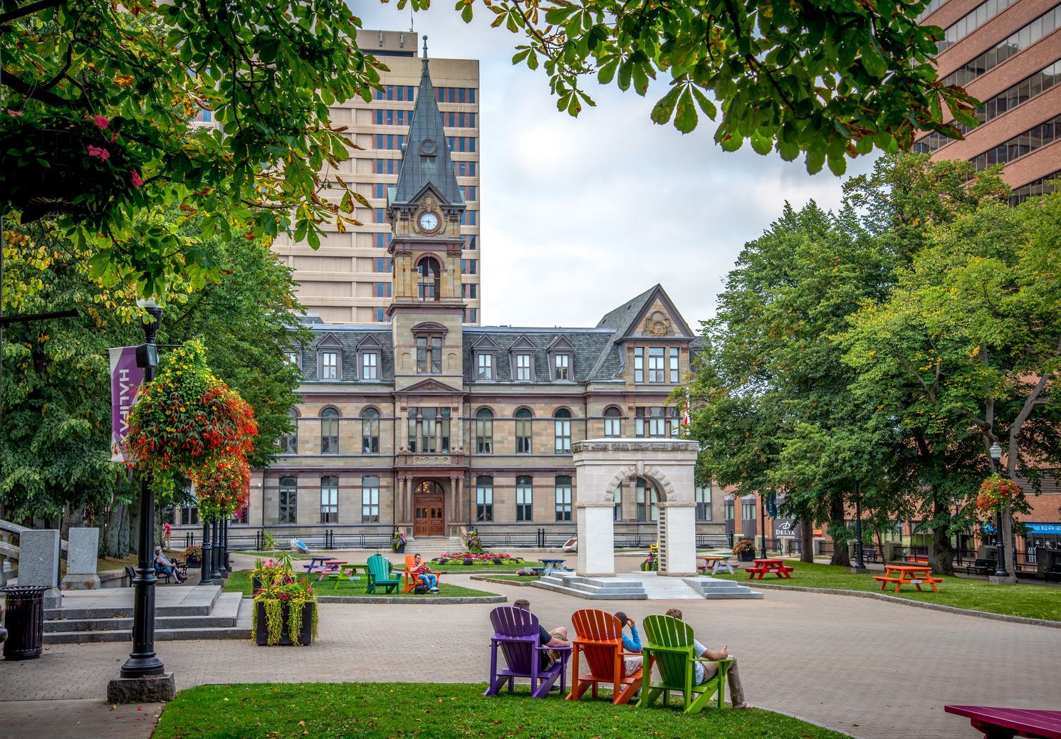 Het stadhuis van  Halifax op Nova Scotia, Canada, omgeven door bomen, met kleurrijke stoelen en bloemperken op een groen openbaar plein.