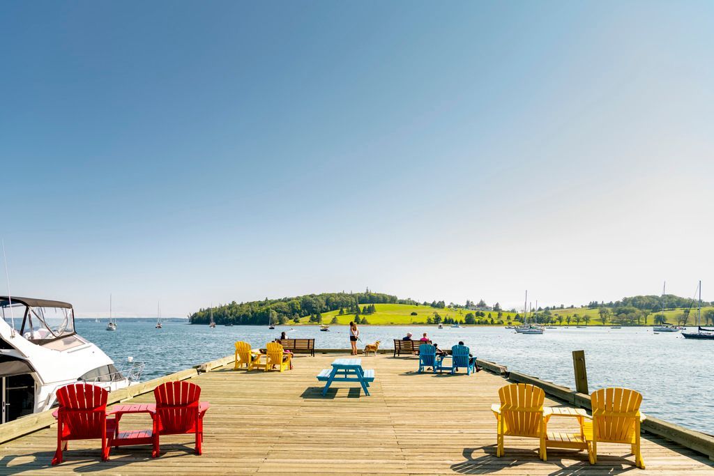 Kleurrijke Adirondack-stoelen op een houten steiger in Lunenburg, Nova Scotia, met uitzicht op een kalm water en een klein groen eilandje onder een heldere hemel.