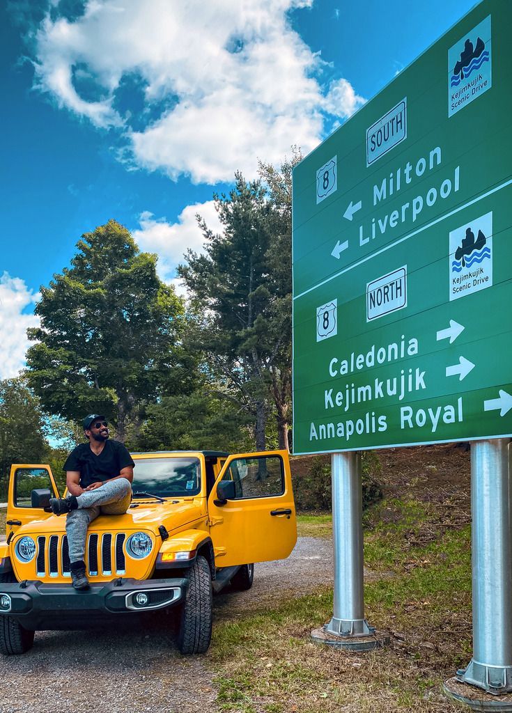 Man in een gele Jeep naast groene verkeersborden op Nova Scotia die wijzen naar Liverpool, Californië en Atlantic Royal onder een blauwe hemel.