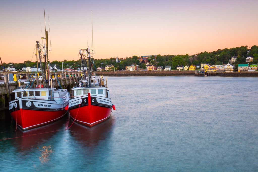 Twee rode vissersboten liggen aangemeerd in een kalme haven van Digby in Nova Scotia bij zonsondergang, met de kustlijn in de verte.