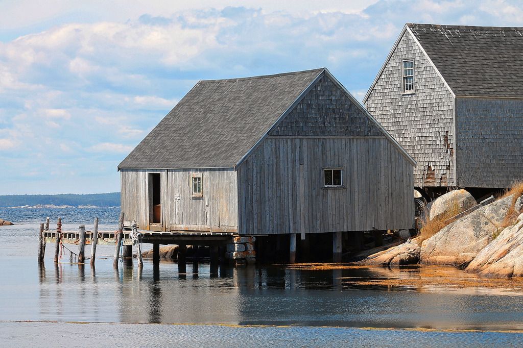 Verweerde houten boothuizen in Peggy's Cove op Nova Scotia op palen aan het kalme water, naast een rotsachtige kust onder een blauwe hemel.
