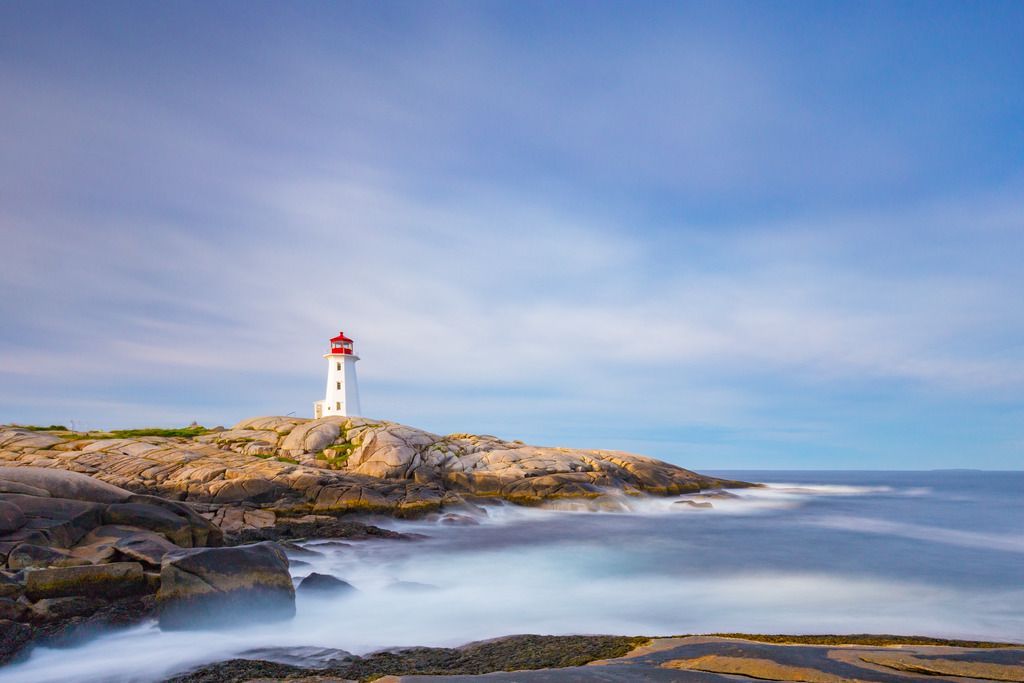 De vuurtoren van Peggy's Cove op Nova Scotia op een rotsachtige kust onder een blauwe hemel, met kalm water op de voorgrond.