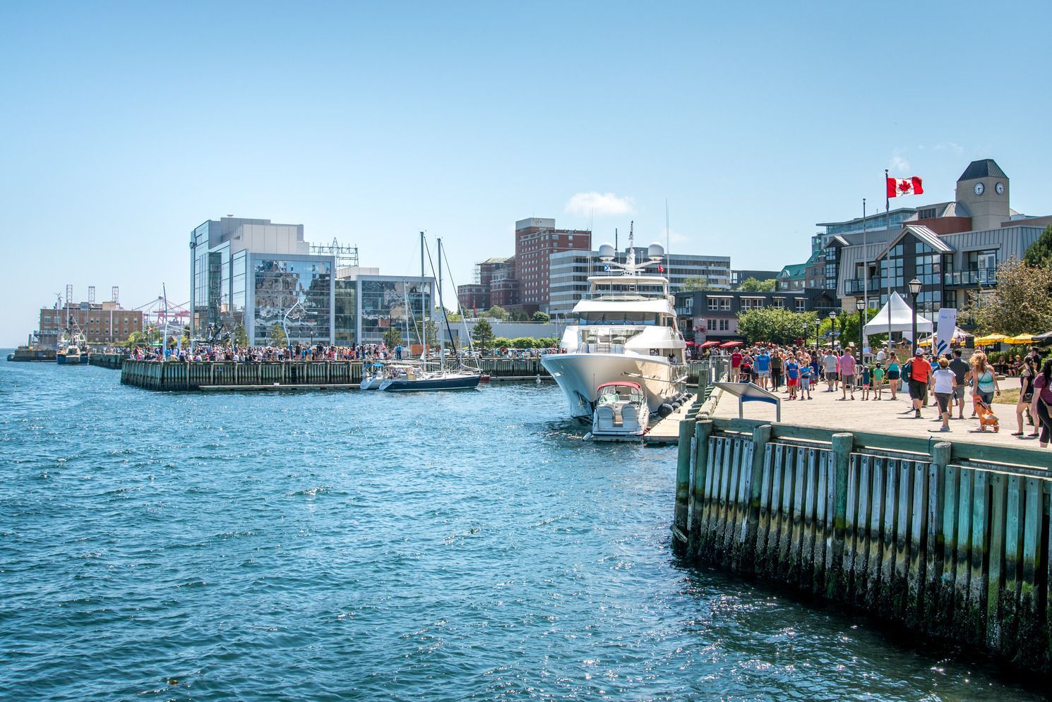 Haven in Halifax op Nova Scotia, Canada, met boten, gebouwen aan het water en mensen die onder een blauwe hemel over een zonnige pier wandelen.