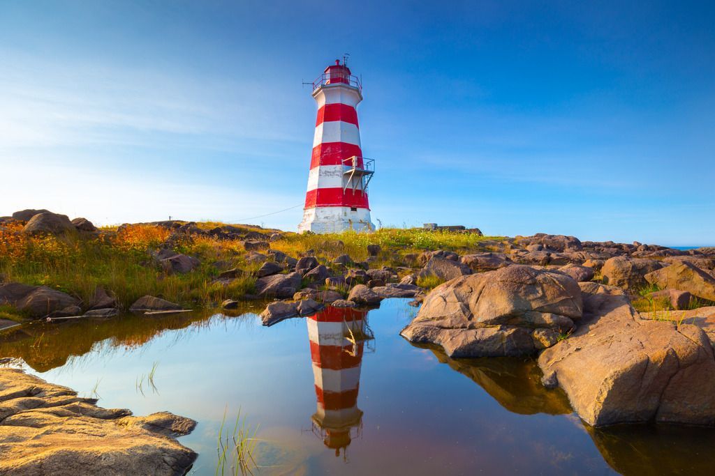 De rood-wit gestreepte vuurtoren Western Light op Brier Island, Nova Scotia, aan een rotsachtige kust met de weerspiegeling van het kalme water tegen een blauwe hemel.