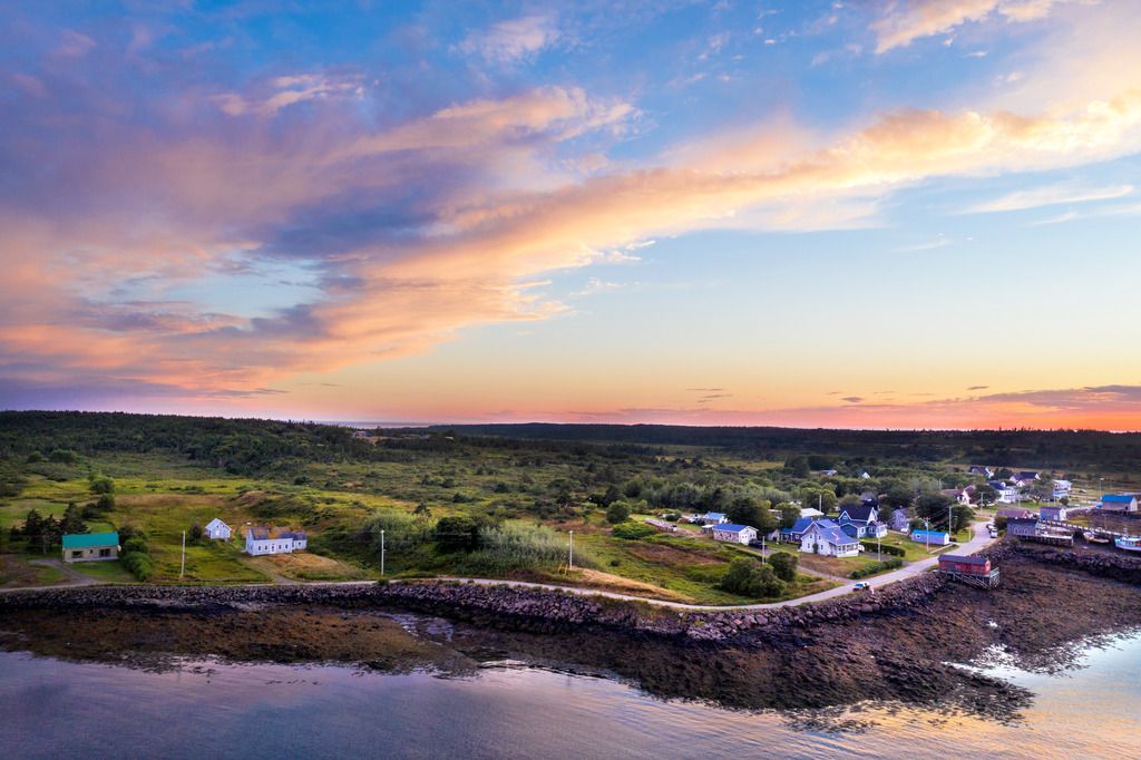 Het kustdorpje Westpoint op Brier Island, Nova Scotia, bij zonsondergang met huizen, een stenen kustlijn en roze-oranje wolken boven kalm water.