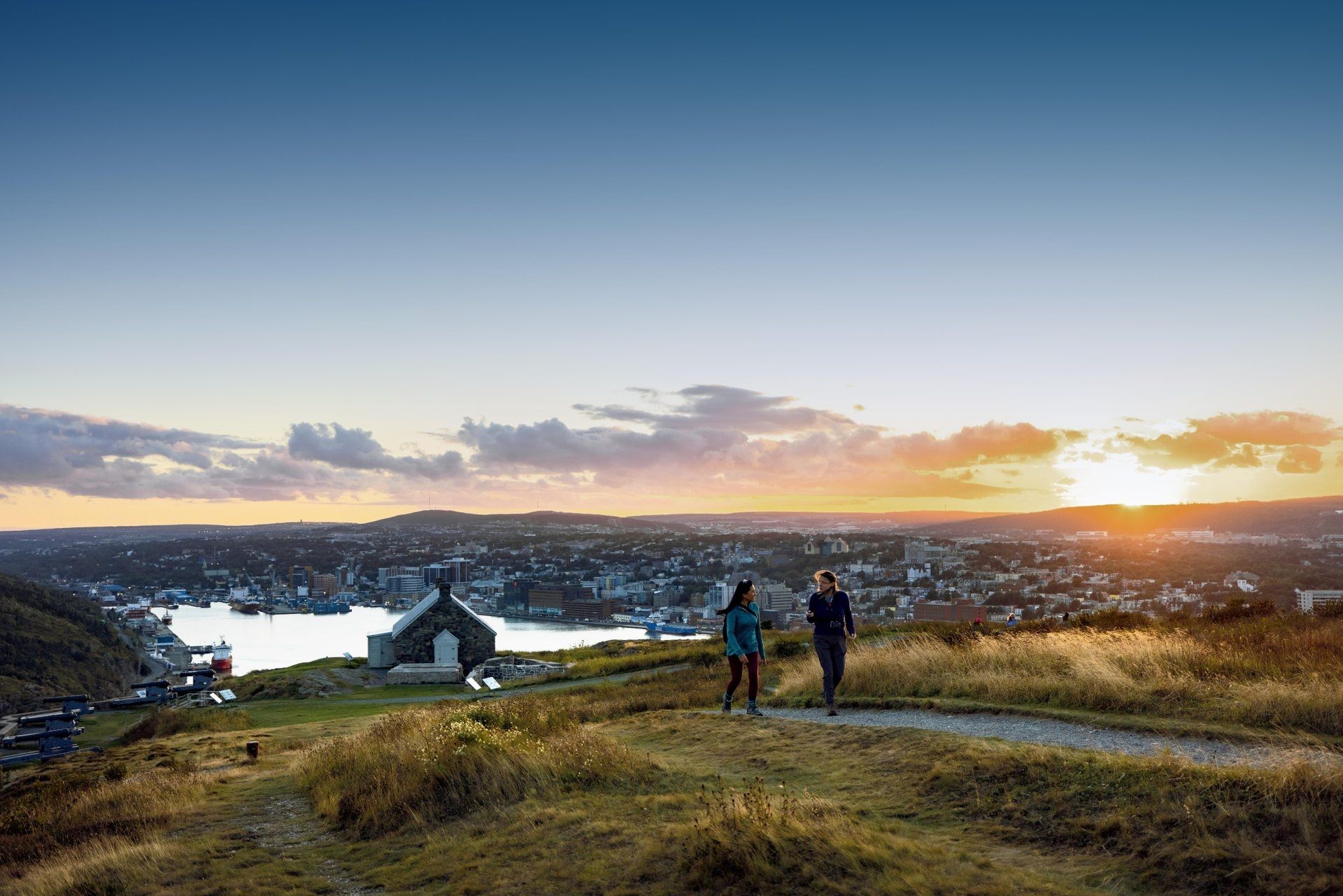 Twee wandelaars op een grasheuvel bij Signal Hill National Historic Site met uitzicht op St. John's en een rivier bij zonsondergang.