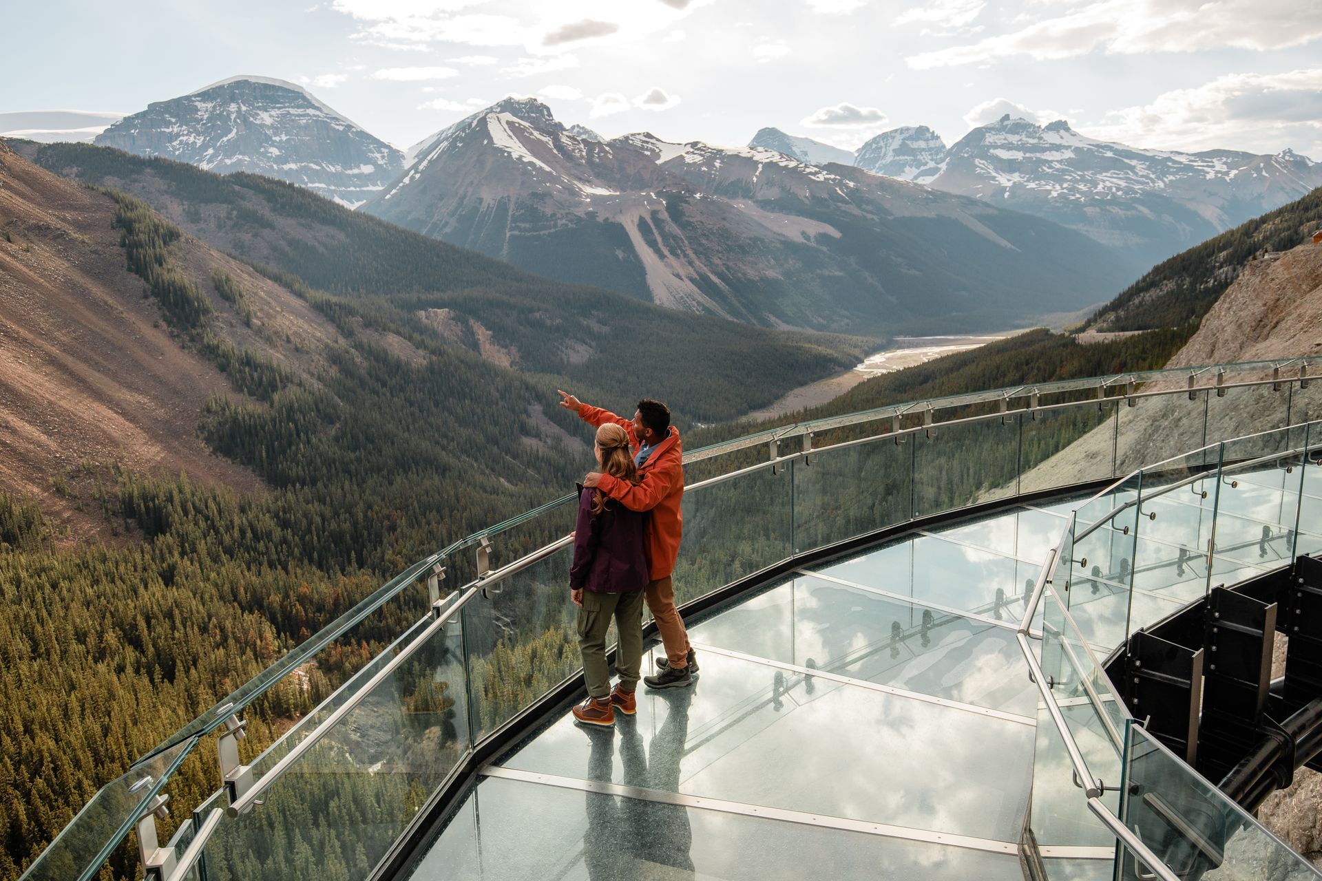 Twee mensen staan ​​op de Skywalk aan de Icefields Parkway en wijzen naar besneeuwde bergen en een beboste vallei.
