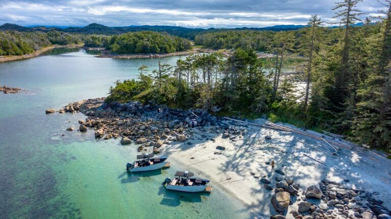 Zandige oever van zee in British Columbia met twee boten, turquoise water, dennenbomen en rotsachtige eilandjes onder een bewolkte hemel.