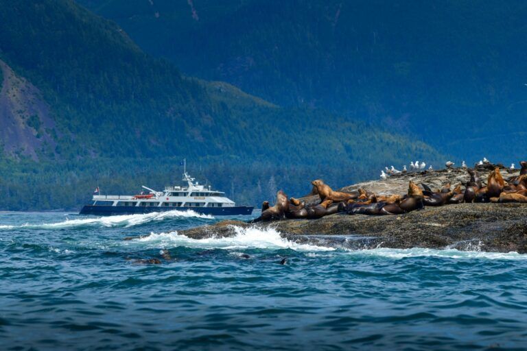 Zandige oever van zee in British Columbia met twee boten, turquoise water, dennenbomen en rotsachtige eilandjes onder een bewolkte hemel.