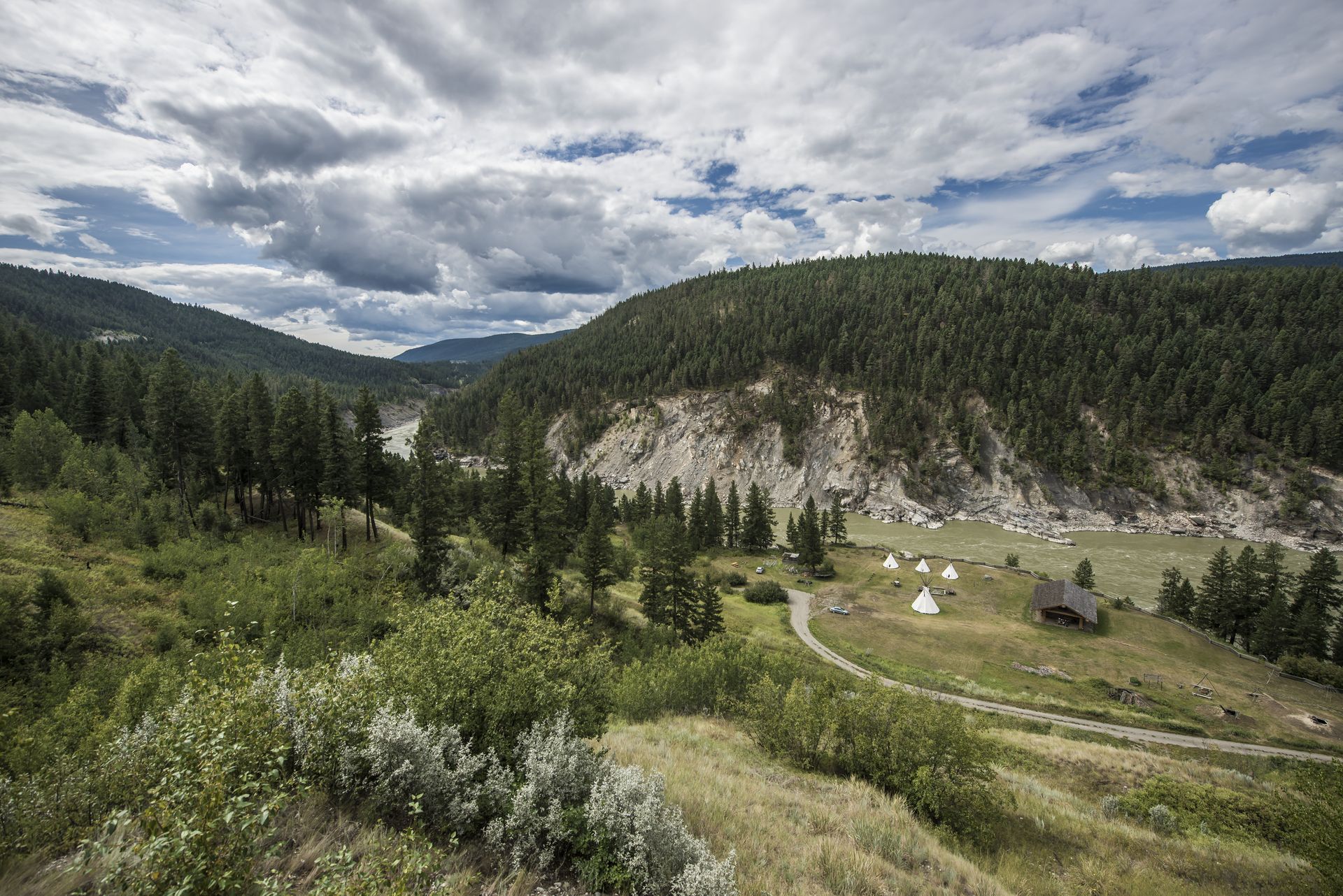 Een brede bergvallei in deFarwell Canyon nabij Riske Creek met beboste heuvels, rotsachtige kliffen, een kronkelende onverharde weg en een bewolkte hemel.