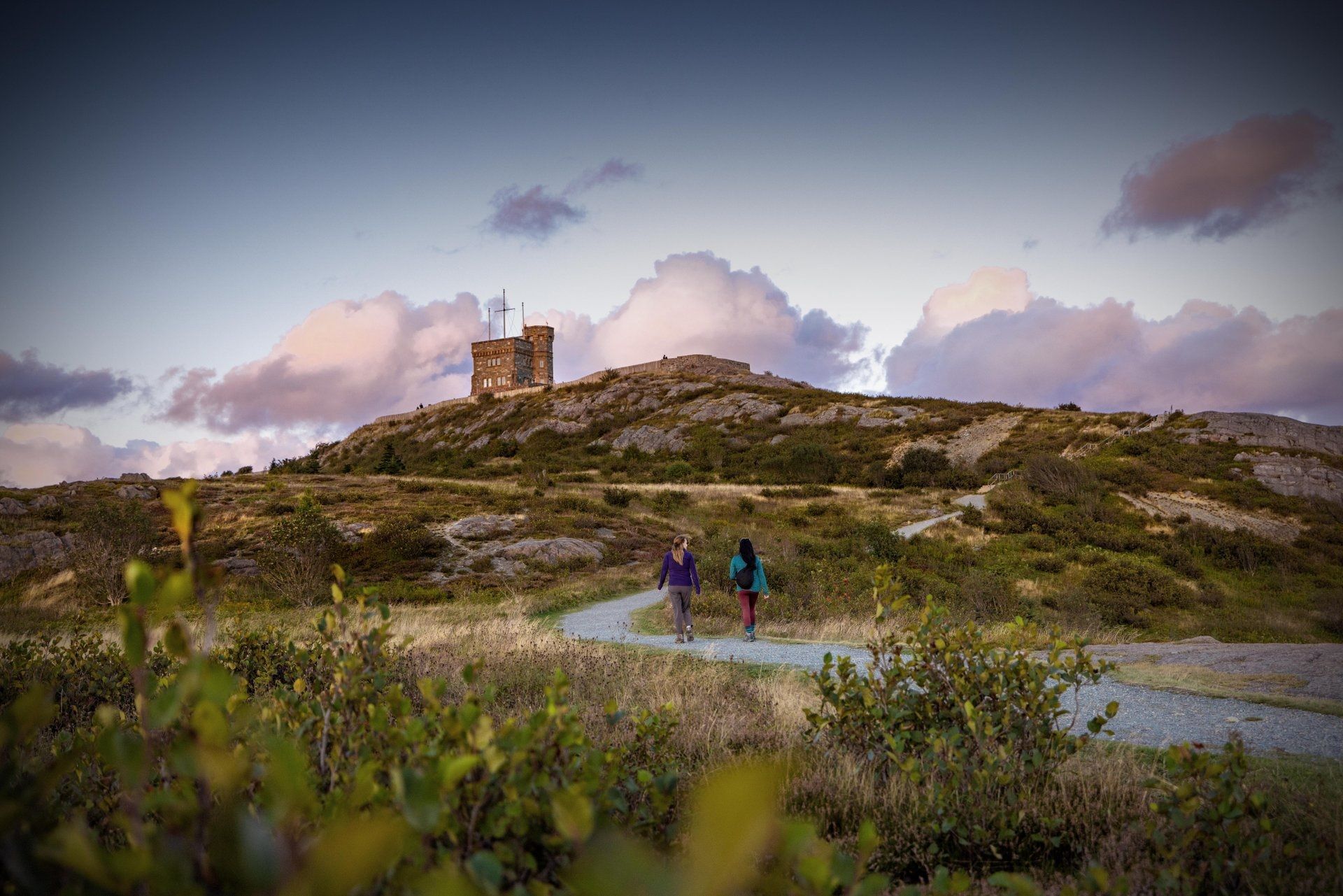 Twee wandelaars op een rotsachtig pad onder een stenen ruïne van Signal Hill in Newfoundland bij zonsondergang.
