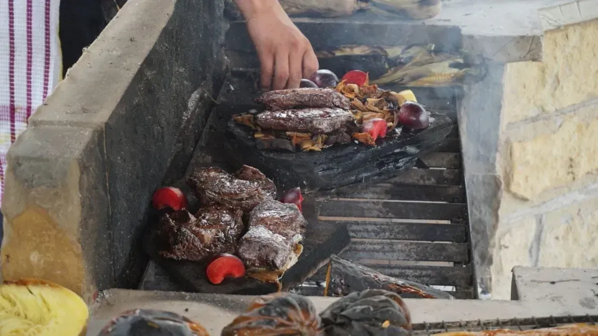 Vlees gegrild boven open kolen in een stenen barbecue, met rook en rode tomaten zichtbaar.