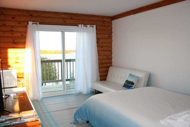 Lichte slaapkamer met wit bed, transparante gordijnen, houten wanden en balkondeuren die uitkomen op de buitenlucht.