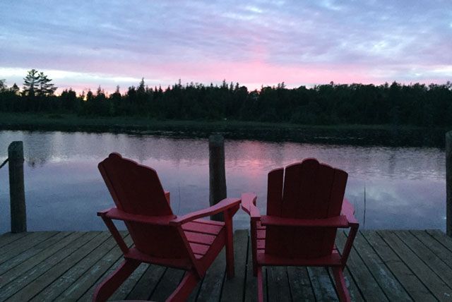 Twee rode Adirondack-stoelen op een steiger met uitzicht op een kalm meer in de schemering.