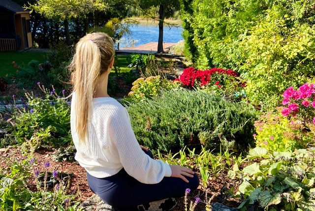 Een vrouw zit in een weelderige tuin en kijkt uit over een zonovergoten blauwe vijver en kleurrijke bloemen.