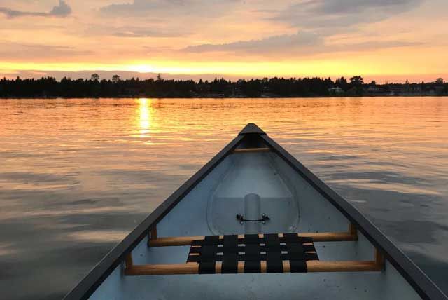 De boeg van een kano op kalm water bij zonsondergang, met gouden reflecties en een roze-oranje lucht.
