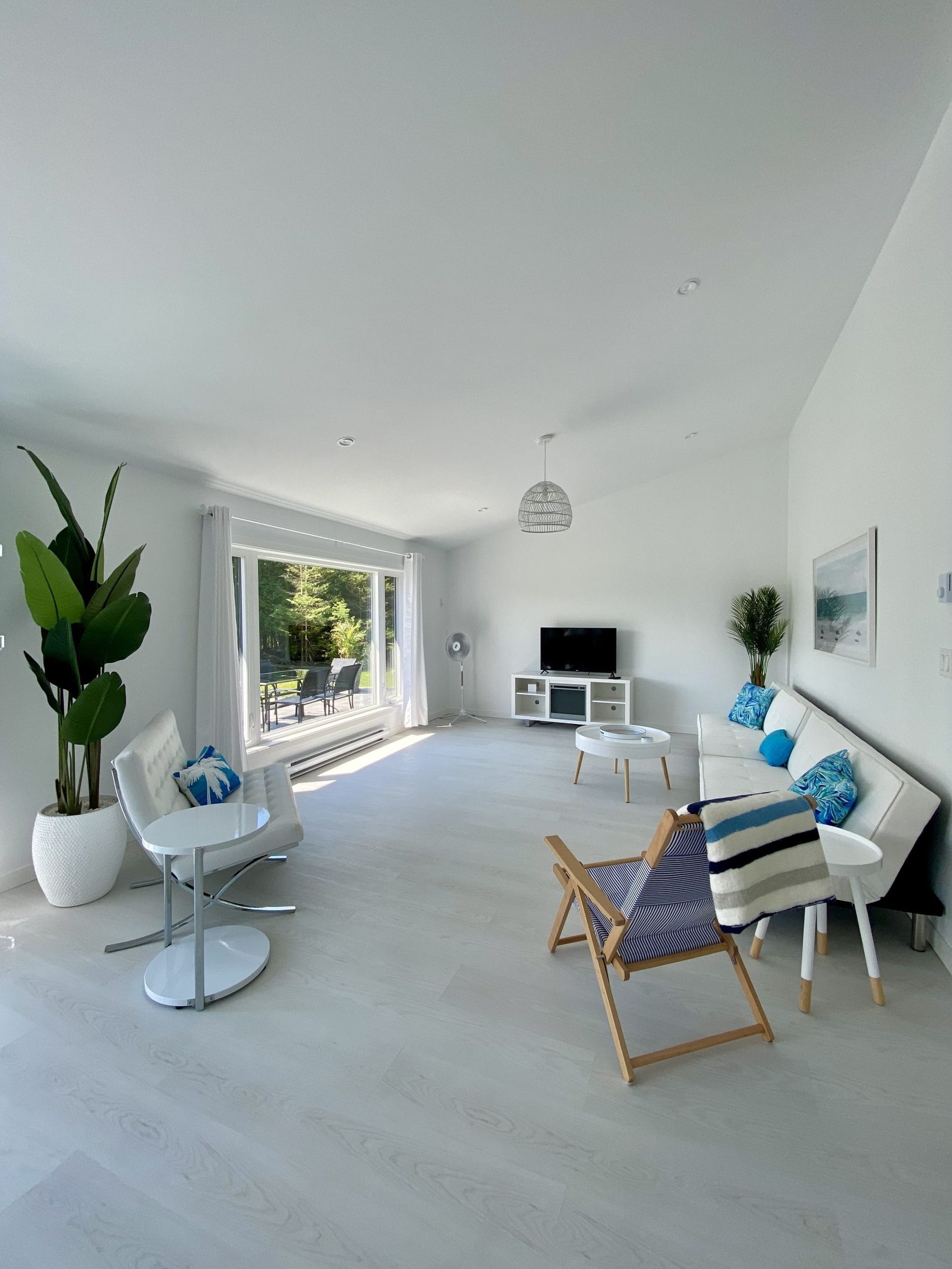 Lichte, minimalistische woonkamer met witte muren, blauwe accenten, groot raam, bank, stoelen en tv-meubel.