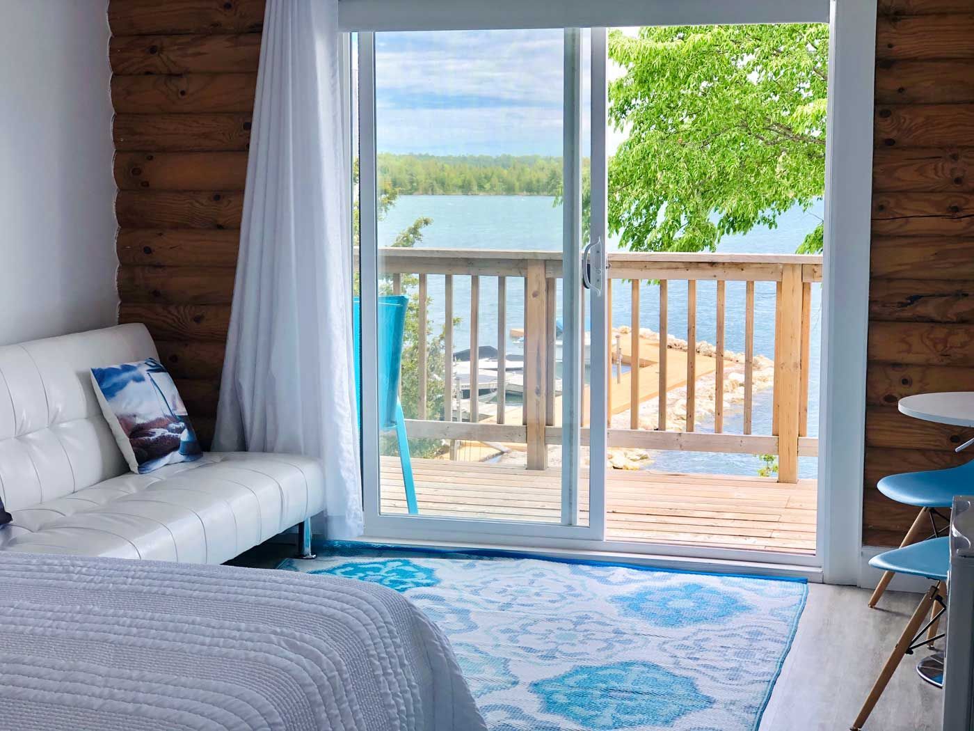 Lichte kamer aan het meer met een witte bank, een blauw vloerkleed en openslaande deuren naar een houten terras met uitzicht op het water.