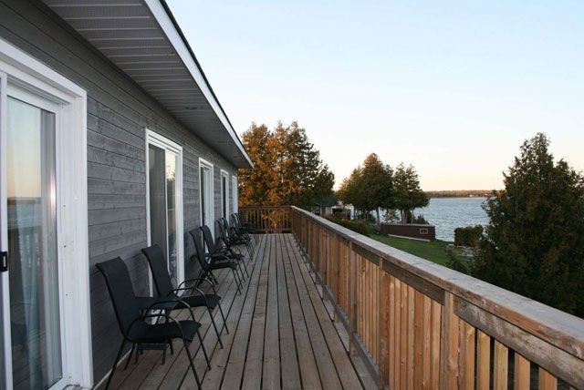 Houten terras naast een grijs huis met uitzicht op het water bij zonsondergang, met zwarte stoelen langs de reling.