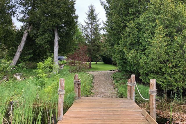 Een houten loopbrug leidt naar een weelderig tuinpad langs een vijver en hoge bomen.