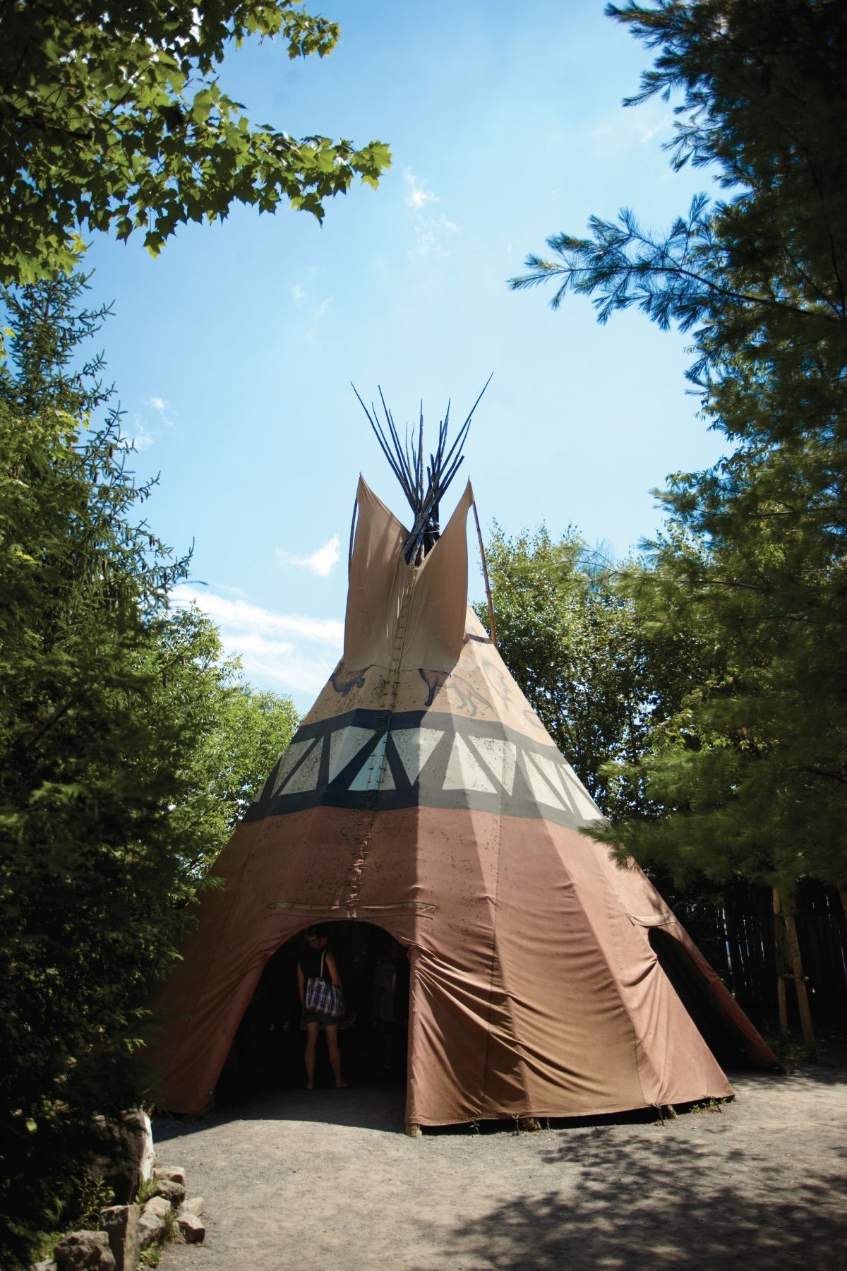 Een beige tipi-tent in Huron Traditional Site ONHOÜA CHETEK8E in Canada met blauwe accenten op een bosrijke camping onder een heldere hemel.