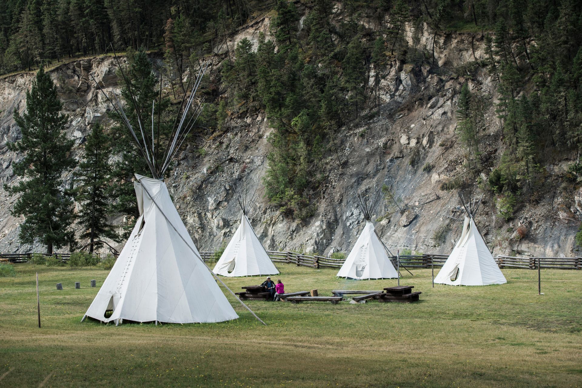 Witte tipi's van Xatsull Heritage Village op een grasveld naast een rotswand, met bomen en een picknicktafel in de buurt.