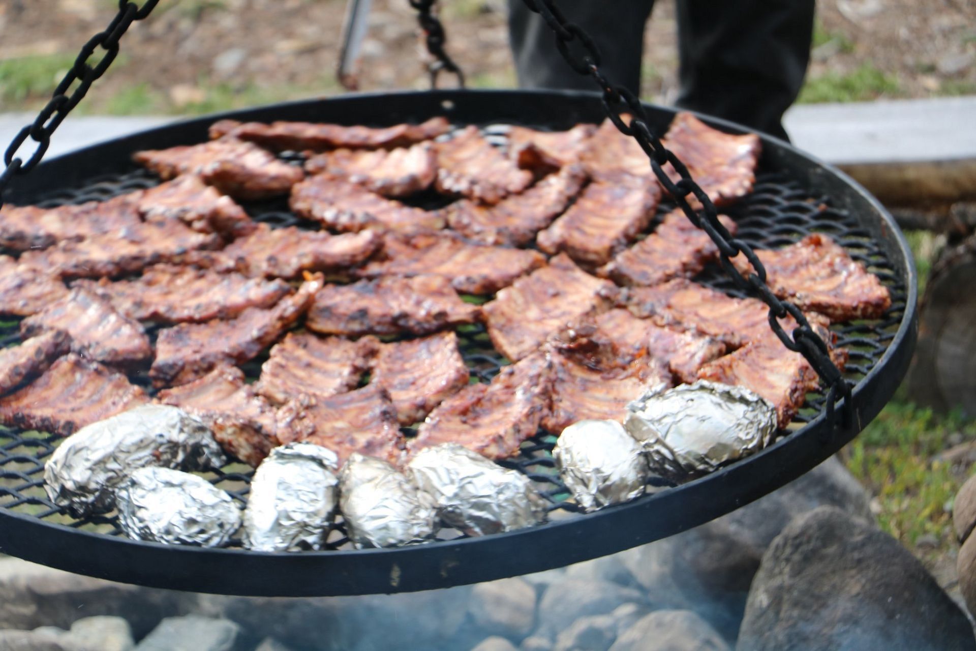 Vlees sist op een grote ronde grill met in folie gewikkelde aardappelen boven een buitenvuur.