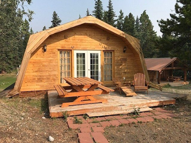 Houten blokhut met een gebogen dak en veranda aan de voorzijde, voorzien van banken en Adirondack-stoelen, in een bosrijke omgeving.
