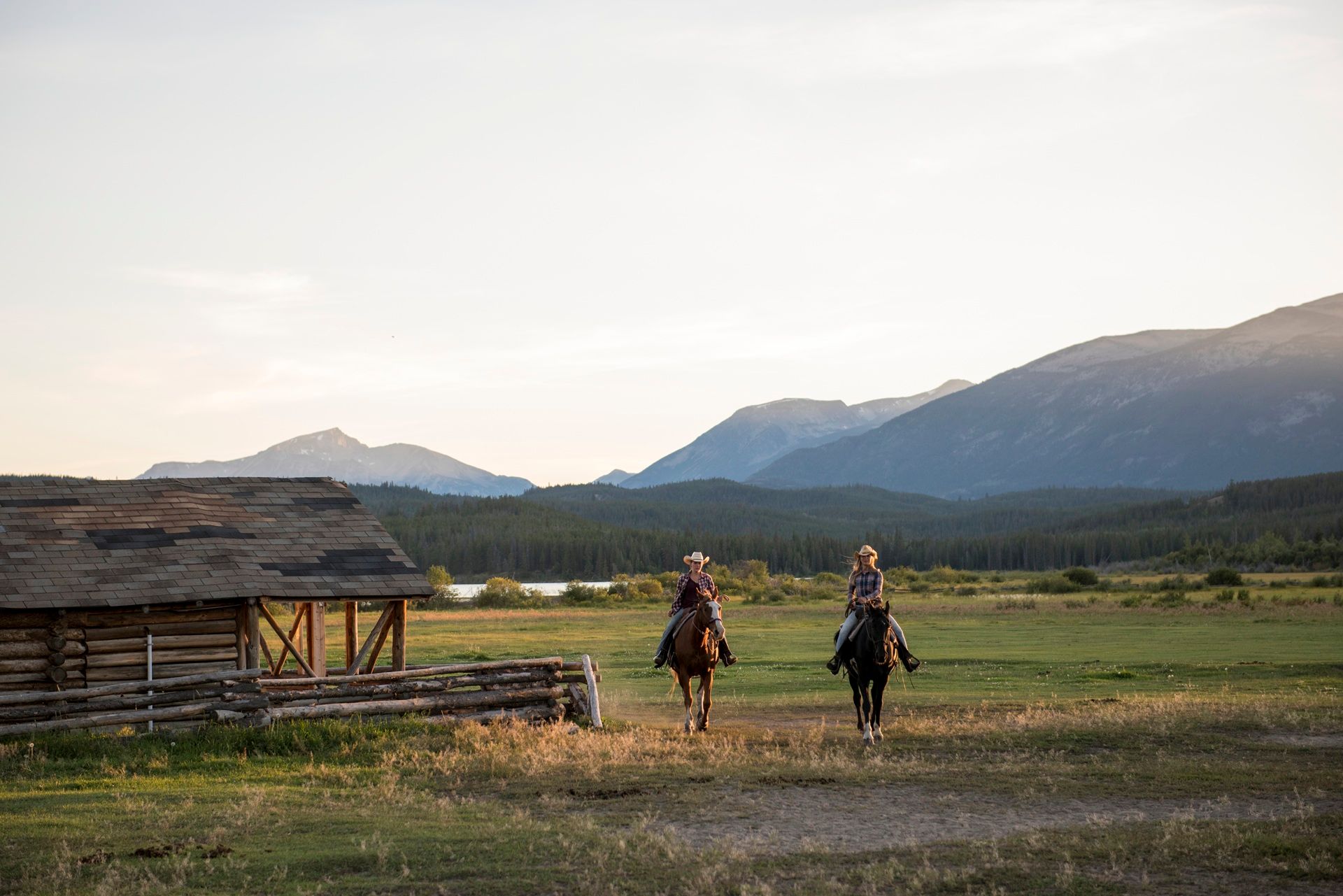Twee ruiters te paard nabij een Terra Nostra Guest Ranch in een grasrijke vallei met bergen op de achtergrond bij zonsondergang.