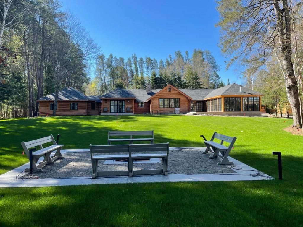 Houten banken en een tafel op een grindterras met uitzicht op een groen gazon en een rustiek huis te midden van hoge bomen.
