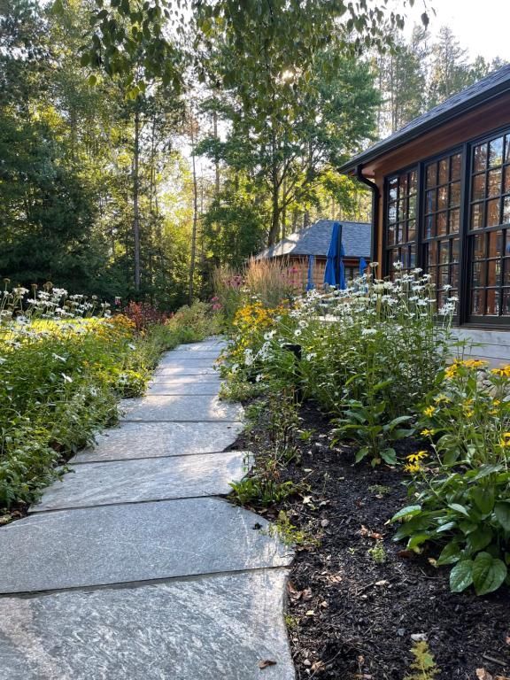 Een stenen tuinpad naast een huis, omzoomd door groene planten en bloemen, leidt naar bomen in het zachte zonlicht.