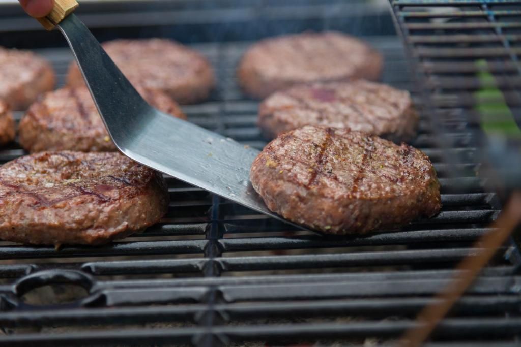Hamburgerpatties worden gegrild op de barbecue, waarbij er eentje met een spatel wordt omgedraaid.