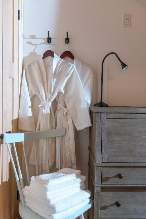 Badkamer met twee witte badjassen aan haken, een stapel witte handdoeken en een grijze commode met een lamp.