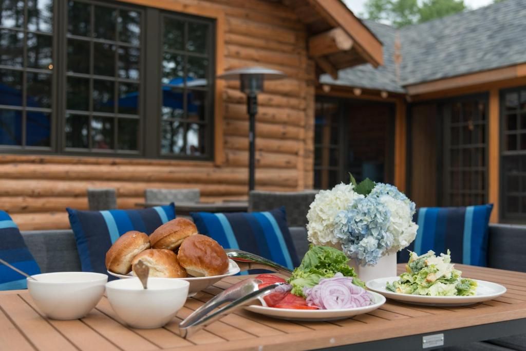 Een buitentafel gedekt met croissants, schalen, salade en bloemen op een terras bij een blokhut, met blauwe stoelen.