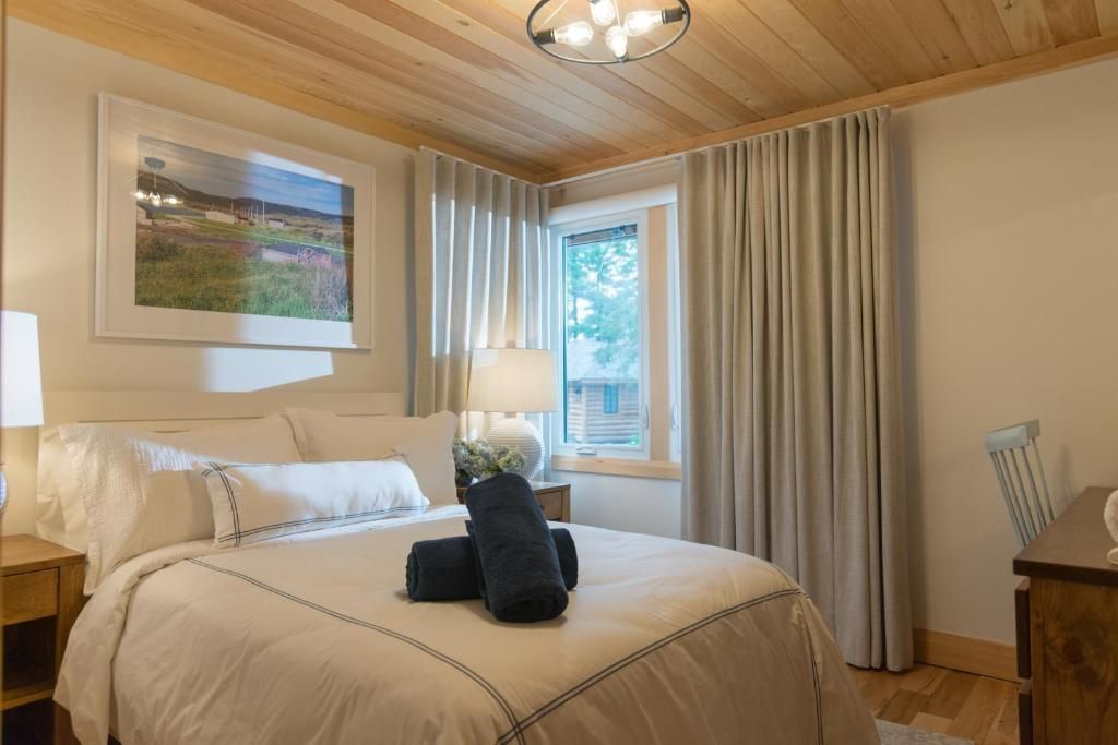 Gezellige slaapkamer met crèmekleurig beddengoed, houten plafond, gestreepte gordijnen en een raam met uitzicht op de tuin.