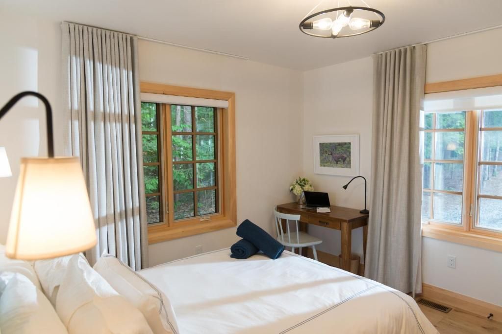 Lichte slaapkamer met wit beddengoed, houten ramen, een lamp en een klein bureau tegen de muur.