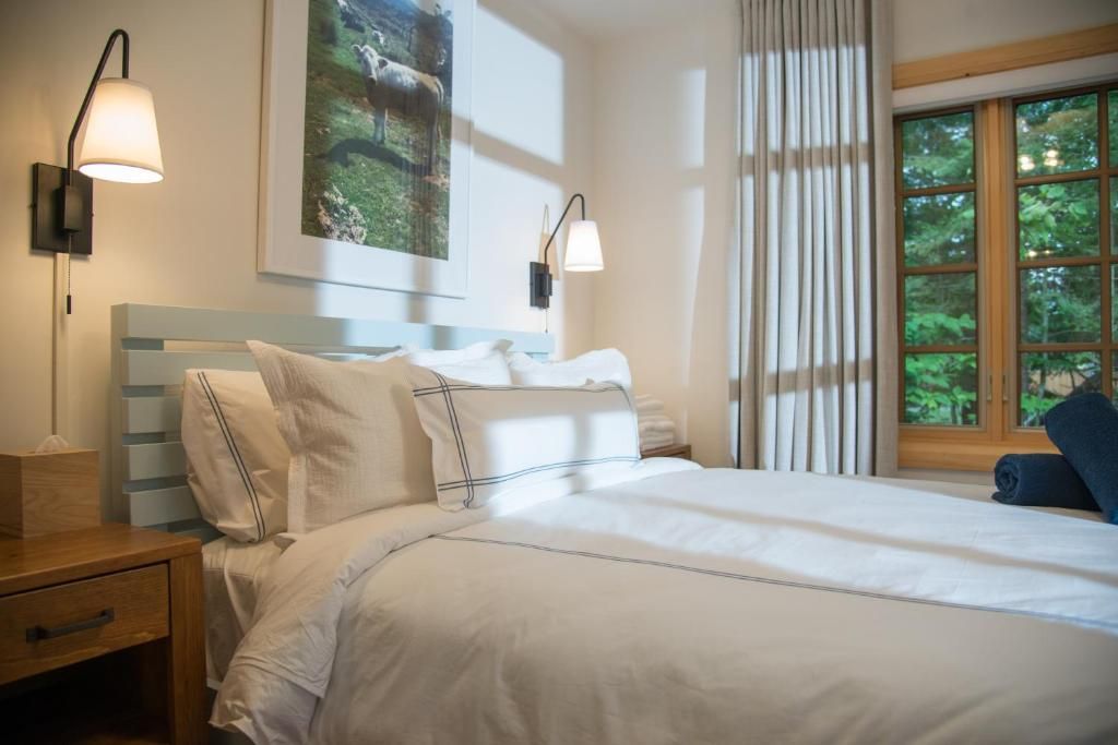 Lichte slaapkamer met een wit bed, nachtlampje, muurdecoratie en een raam met uitzicht op groen.