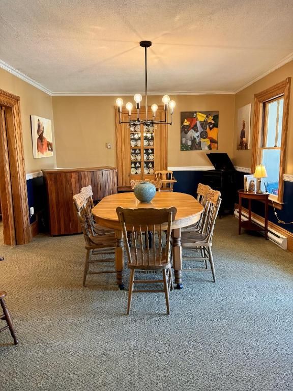 Eetkamer met ronde houten tafel, stoelen, kroonluchter, piano en grote ramen.