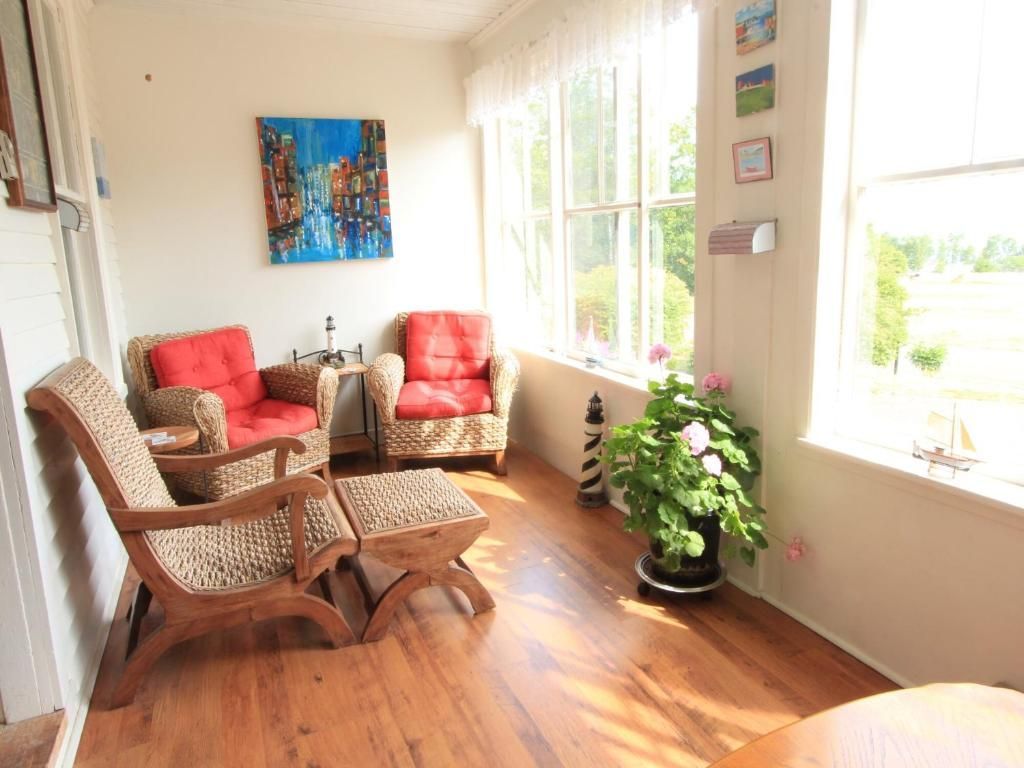 Lichte, zonnige woonkamer met rieten stoelen, rode kussens, planten en grote ramen.