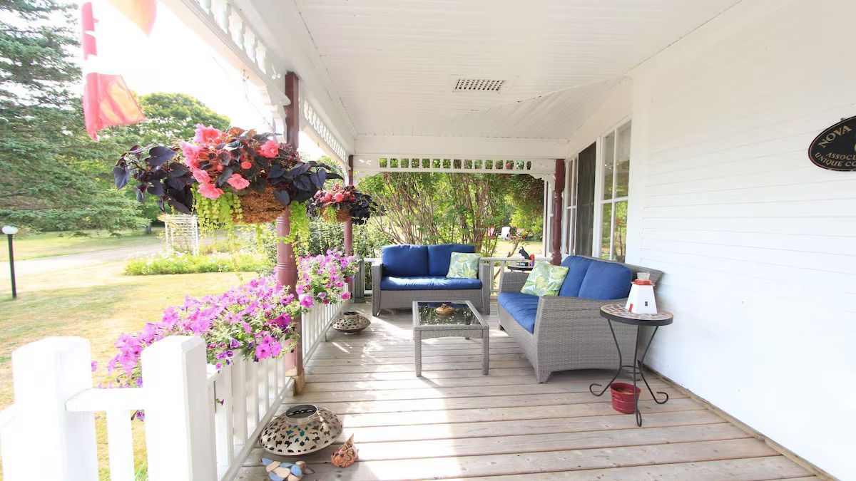 Overdekte veranda aan de voorzijde met blauwe zitmeubels, hangende bloemen en witte balustrade, met uitzicht op een groene tuin.