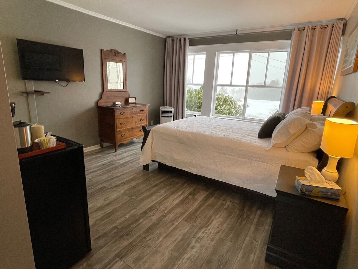 Nette slaapkamer met een bed, commode, aan de muur gemonteerde tv en een groot raam met gordijnen.