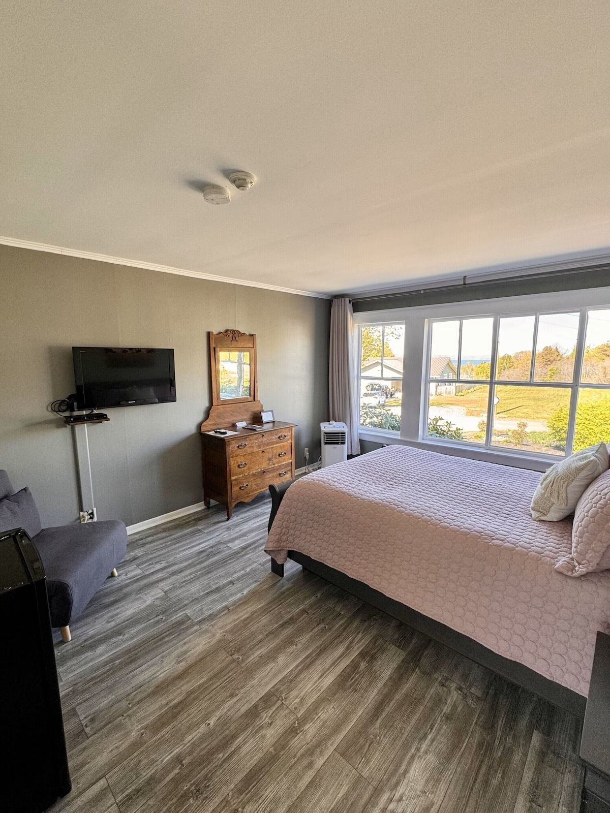 Lichte slaapkamer met een bed, commode, aan de muur gemonteerde tv en grote ramen met uitzicht op herfstbomen.